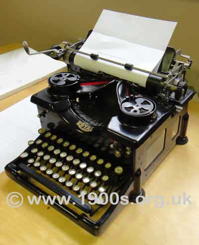 Vintage manual typewriter showing: the rollers holding the paper, the knob to wind the paper through the rollers, the lever to bring the rollers/paper back to position at the end of each line of typing, and the spools taking the two-colour typewriter ribbon